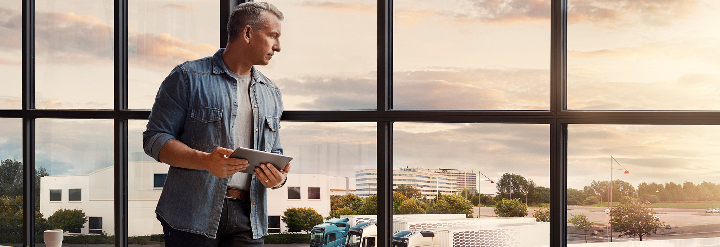 Un uomo con in mano un tablet si trova vicino a una finestra e guarda la sua flotta di camion