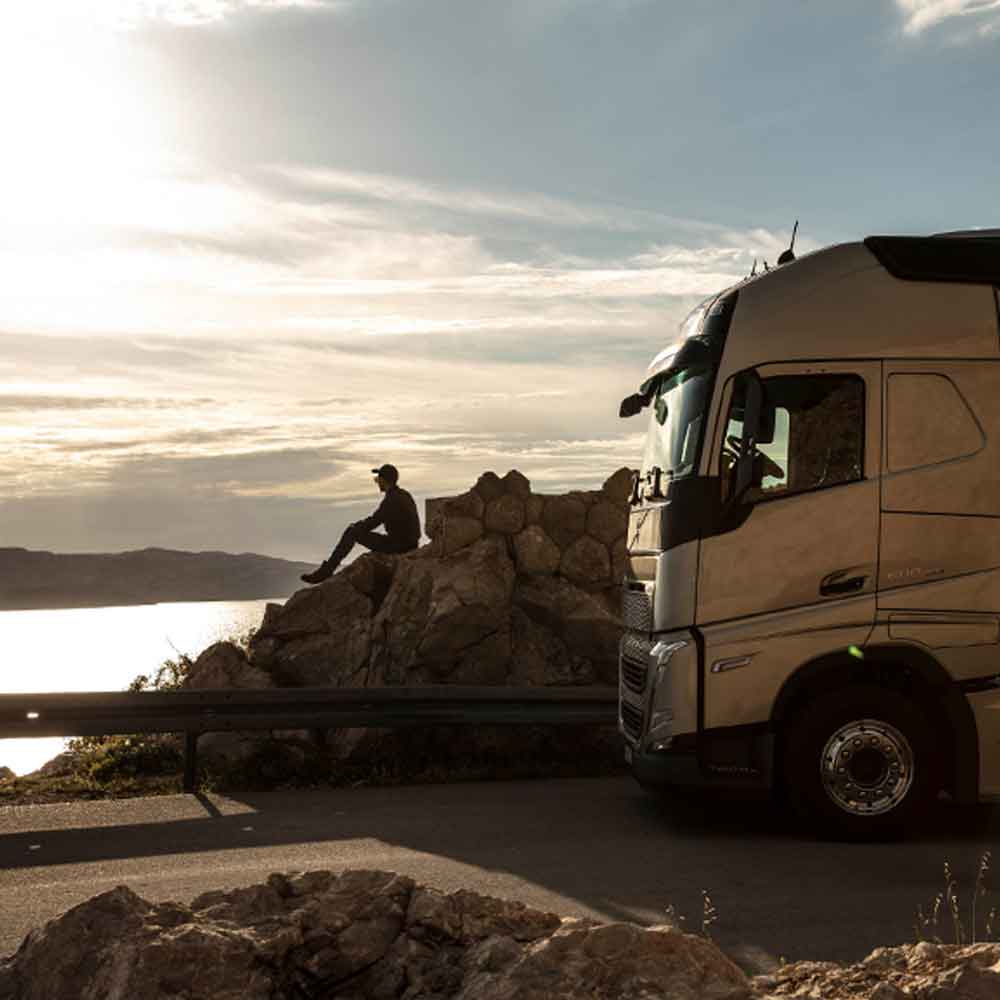 Conducente seduto su una roccia a guardare il mare al tramonto, con il suo veicolo Volvo FH parcheggiato accanto